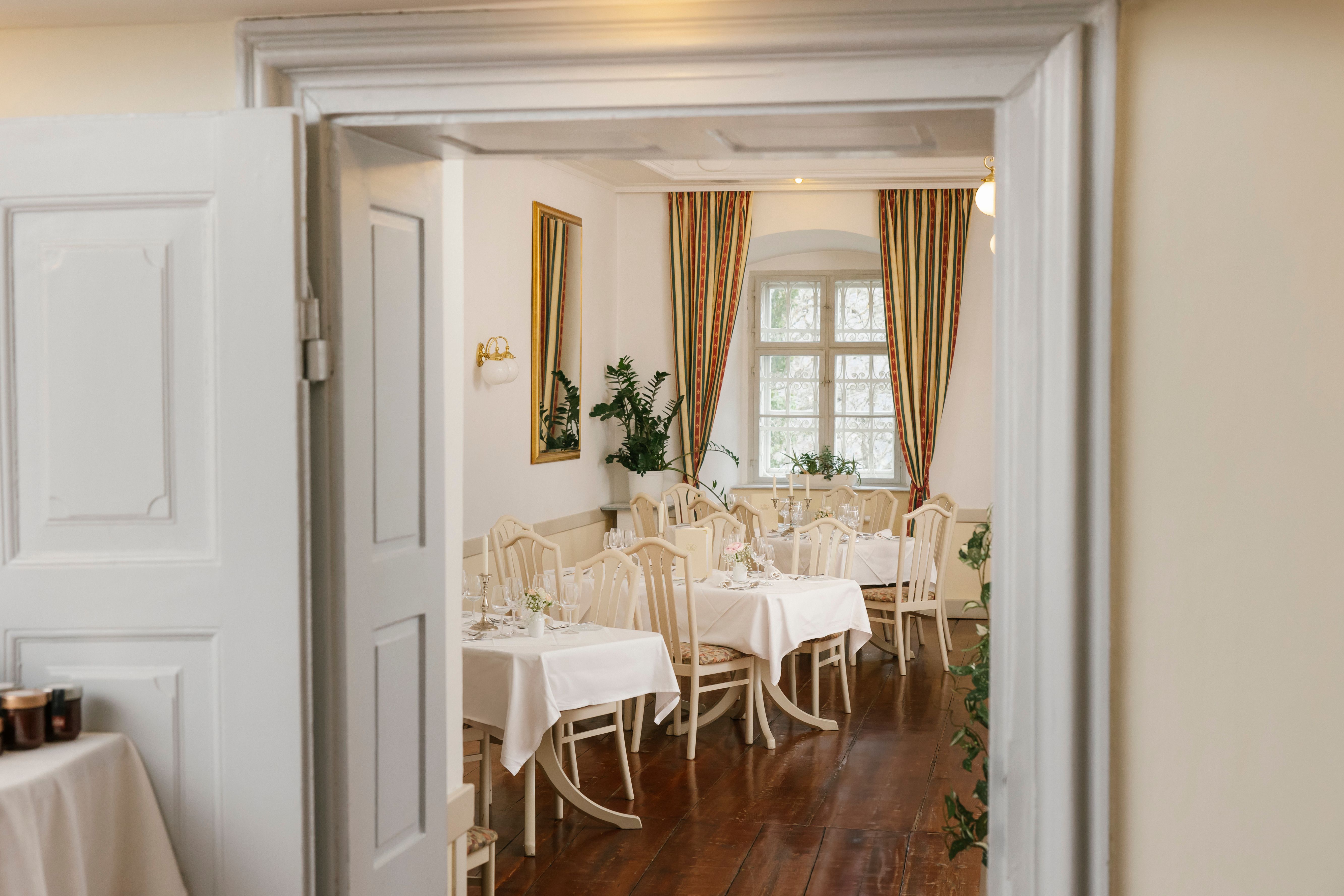 Elegantní restaurace s prostřenými stoly, bílými ubrusy a klasickými židlemi, do které je vidět otevřenými dveřmi.