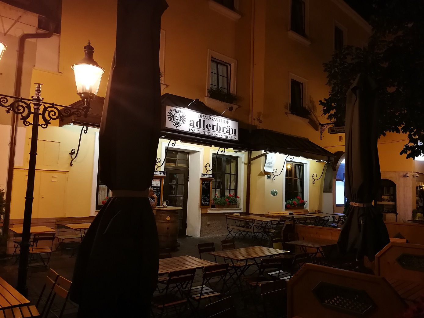 Noční záběr na restauraci s nápisem "Adlerbräu" a prázdnými stoly venku.