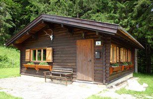 Malý rustikální dřevěný lovecký zámeček s okenicemi a parožím na fasádě, obklopený lesem.