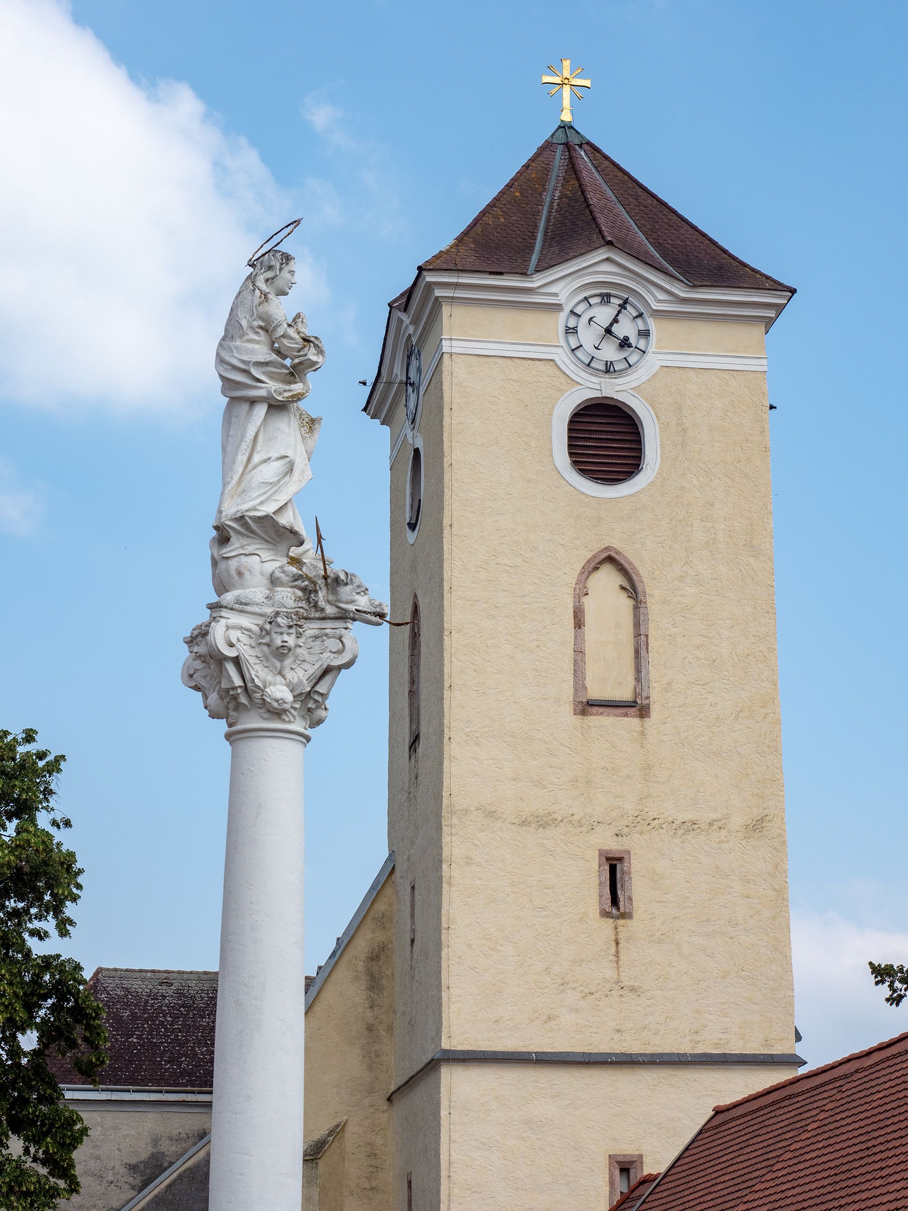 Kostelní věž s hodinami a mariánským sloupem ve Schrattenthalu na pozadí modré oblohy.