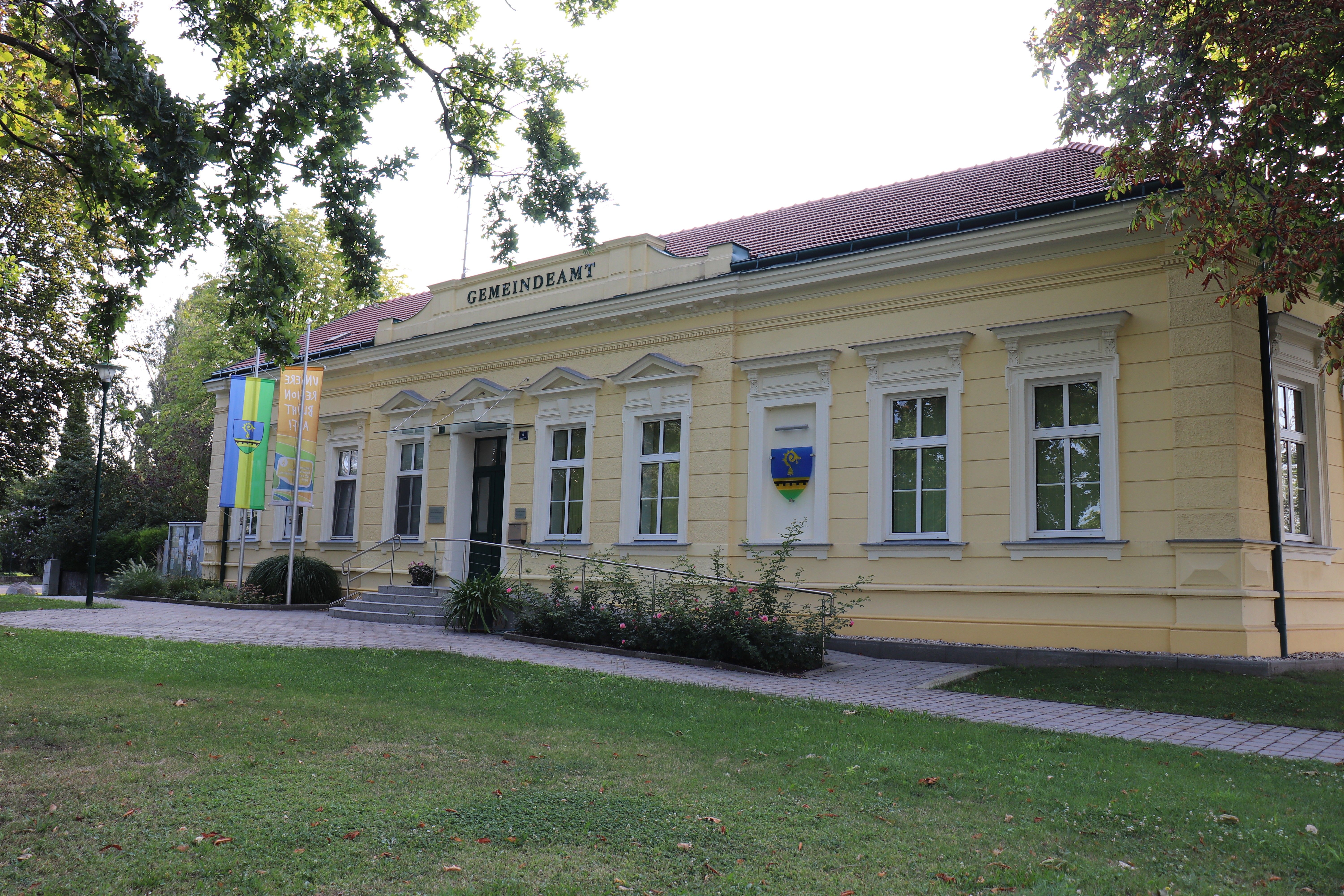 Obecní úřad v Raasdorfu, světlá administrativní budova s červenou střechou, vlajkami a erbem, obklopená stromy a trávníkem.