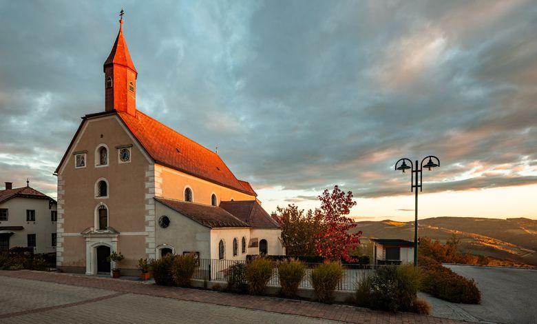 Kostel v St. Corona am Wechsel při západu slunce se zataženou oblohou.