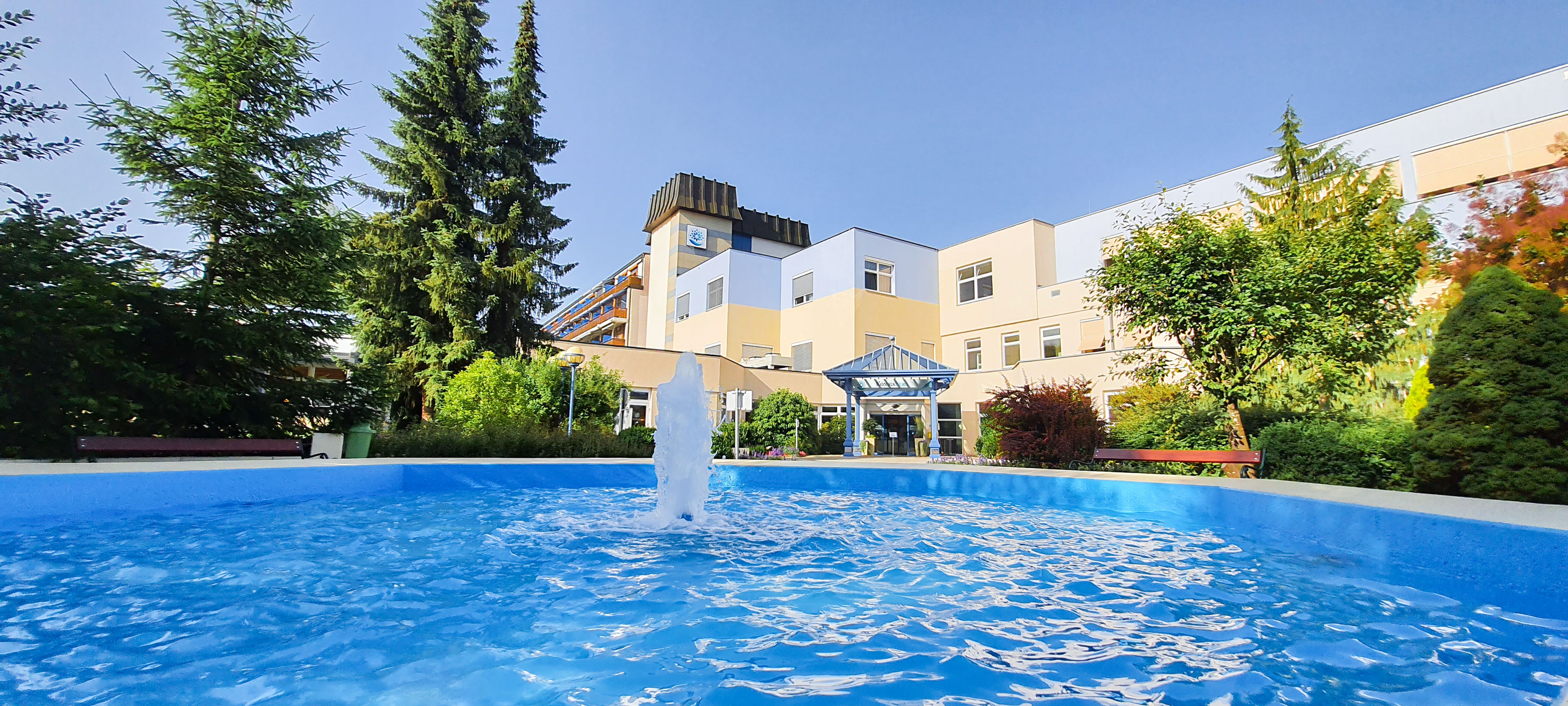 Pohled zvenčí na lázně s fontánou a zahradou.