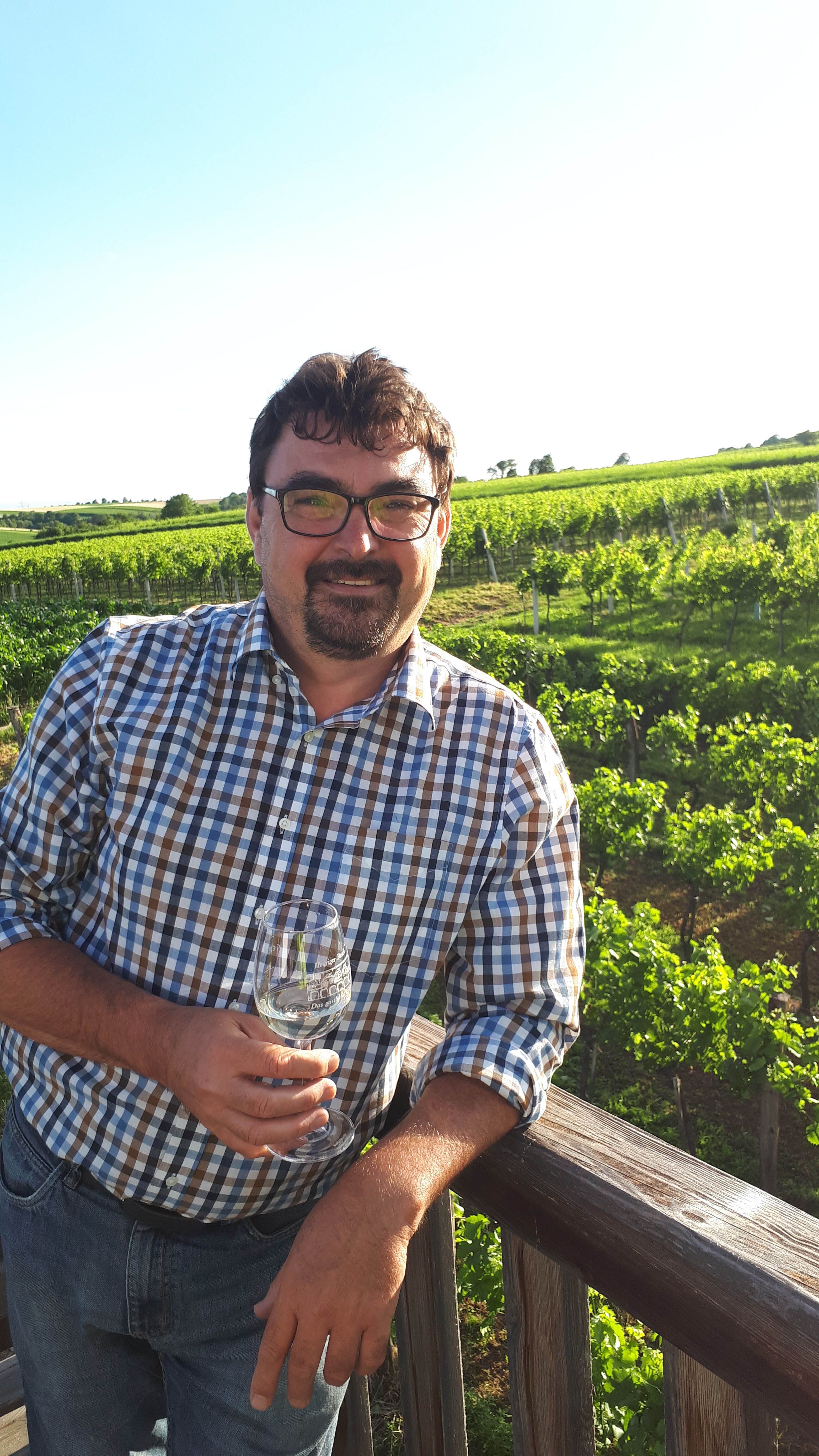 Muž v kostkované košili stojí před vinicí se sklenicí vína v ruce.