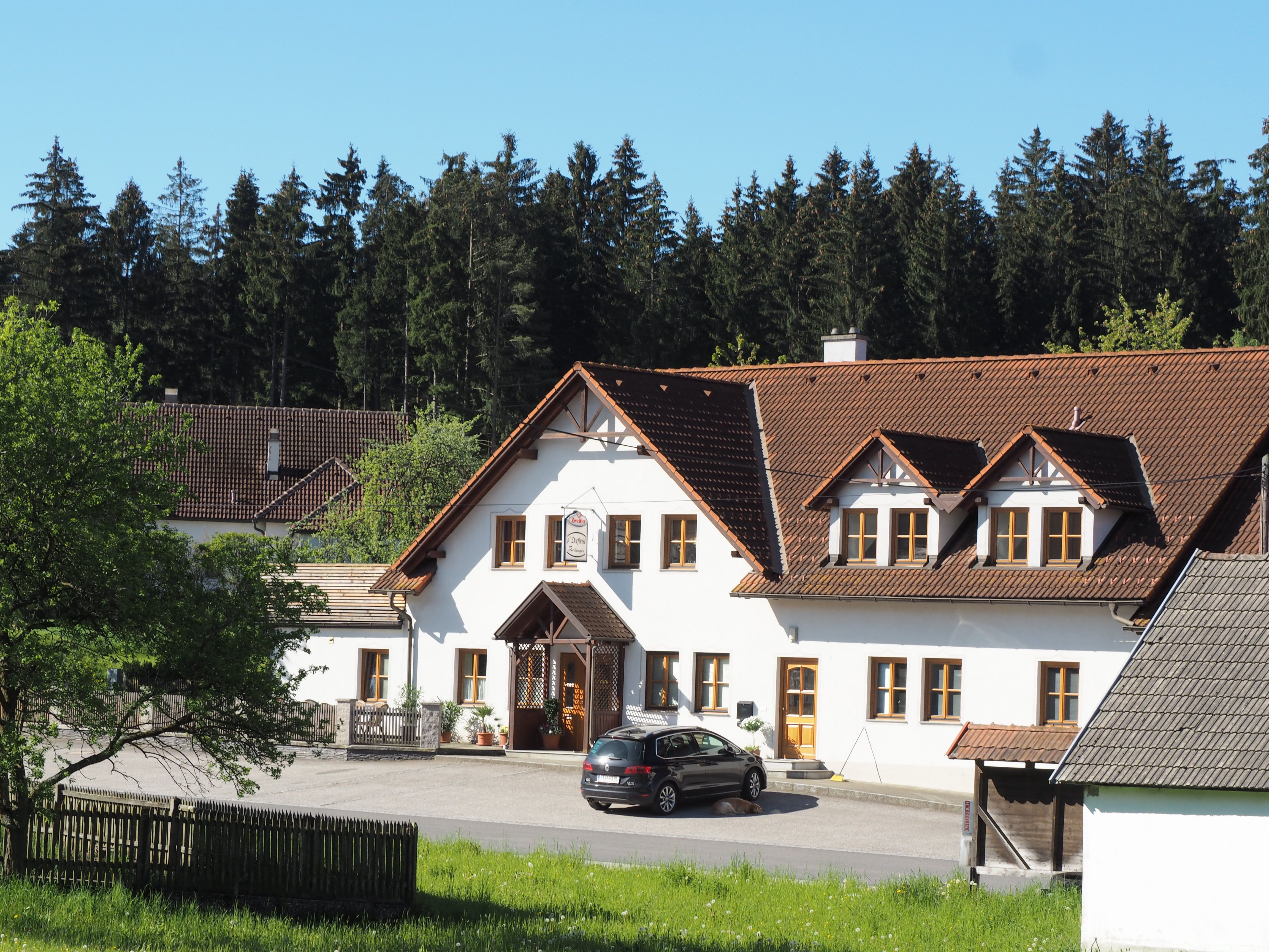 Tradiční hostinec s bílou fasádou a hnědou střechou, obklopený stromy a loukami.