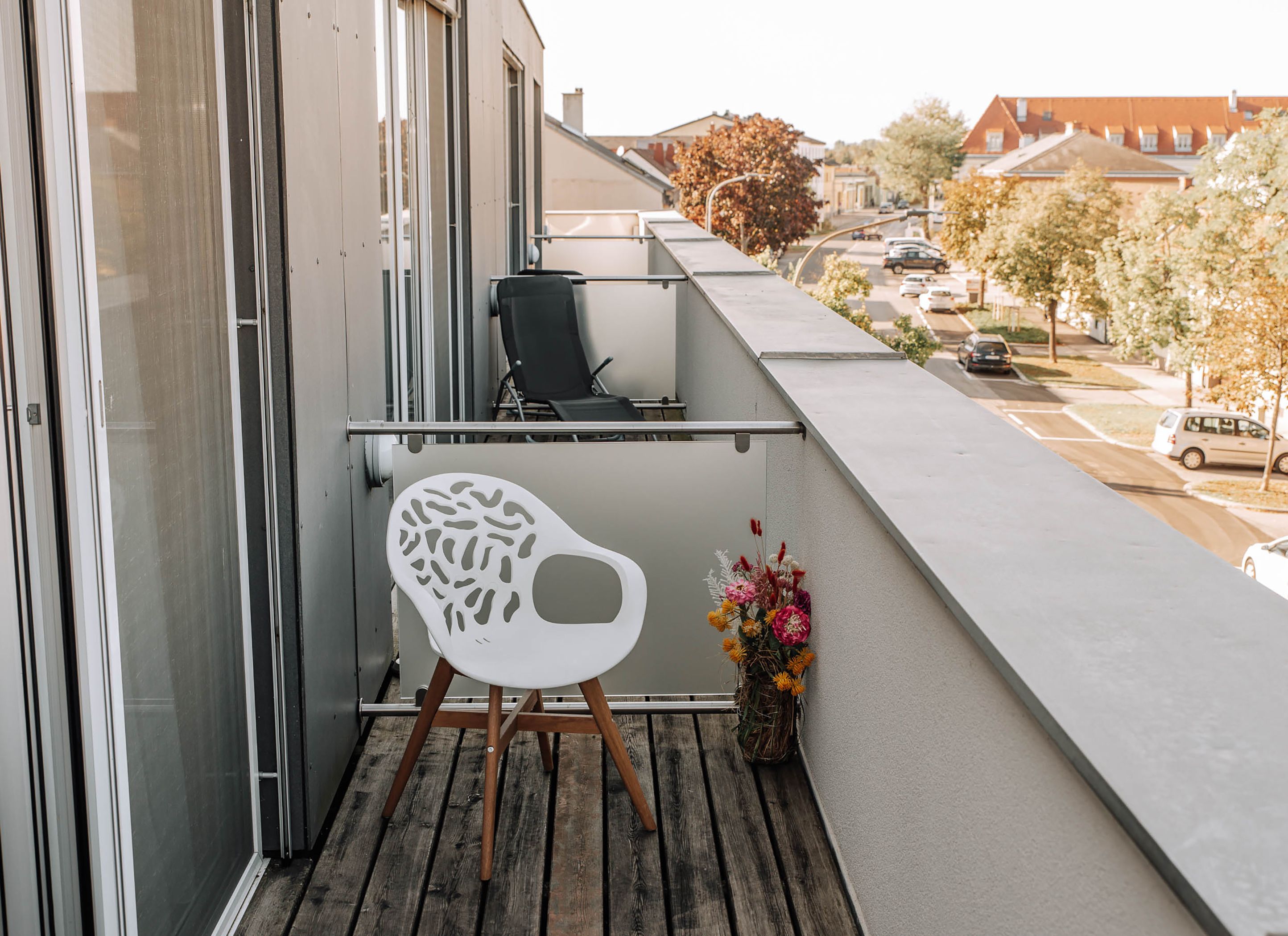 Balkon s bílým křeslem a kyticí květin, výhled na ulici a budovu.