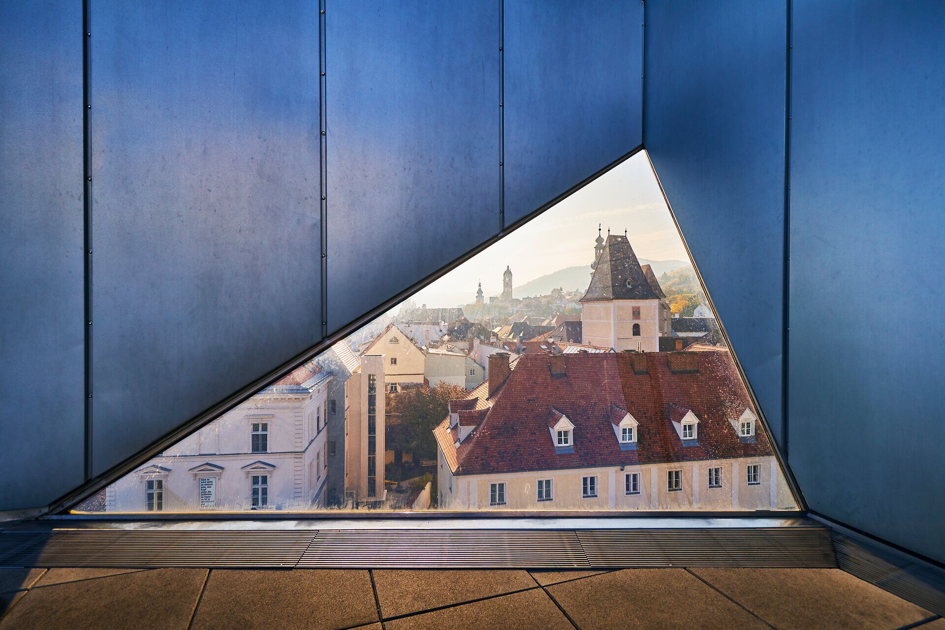 Durch das große, dreieckige Fenster eröffnet sich ein atemberaubender Blick auf die charmante Altstadt mit ihren historischen Gebäuden und dem sanften Hügelpanorama im Hintergrund. Die warmen Farben des Sonnenlichts tauchen die Szene in eine einladende Atmosphäre, die zum Verweilen und Entdecken einlädt.