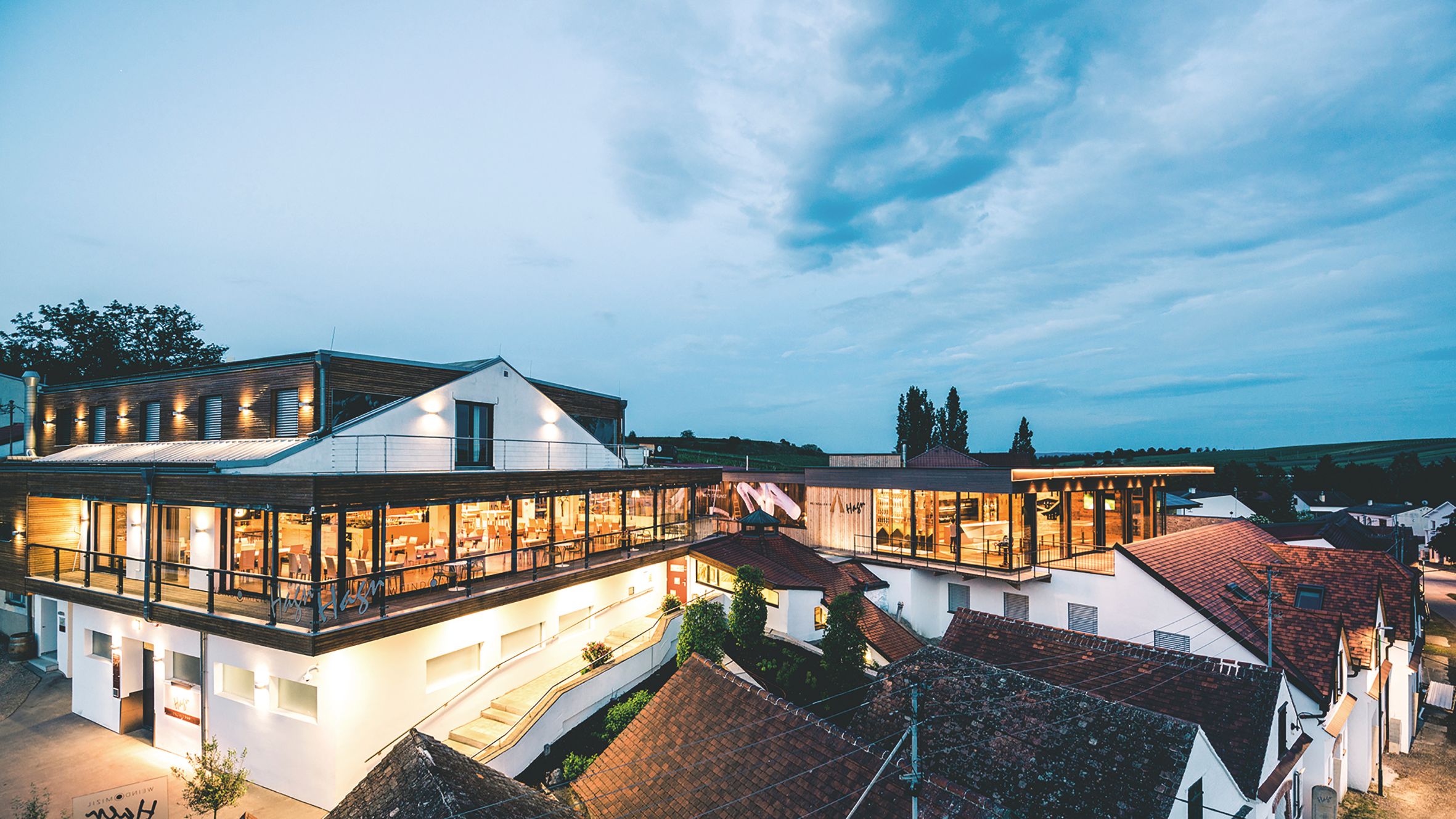 Moderní architektura vinařství za soumraku s osvětlenými interiéry a okolními budovami.