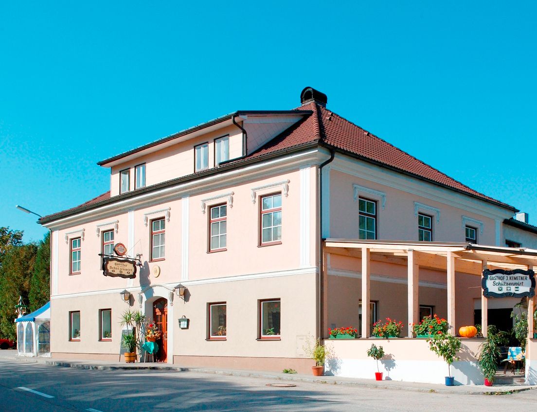 Tradiční hostinec s růžovou fasádou a červenou střechou, zdobený rostlinami a dýní.