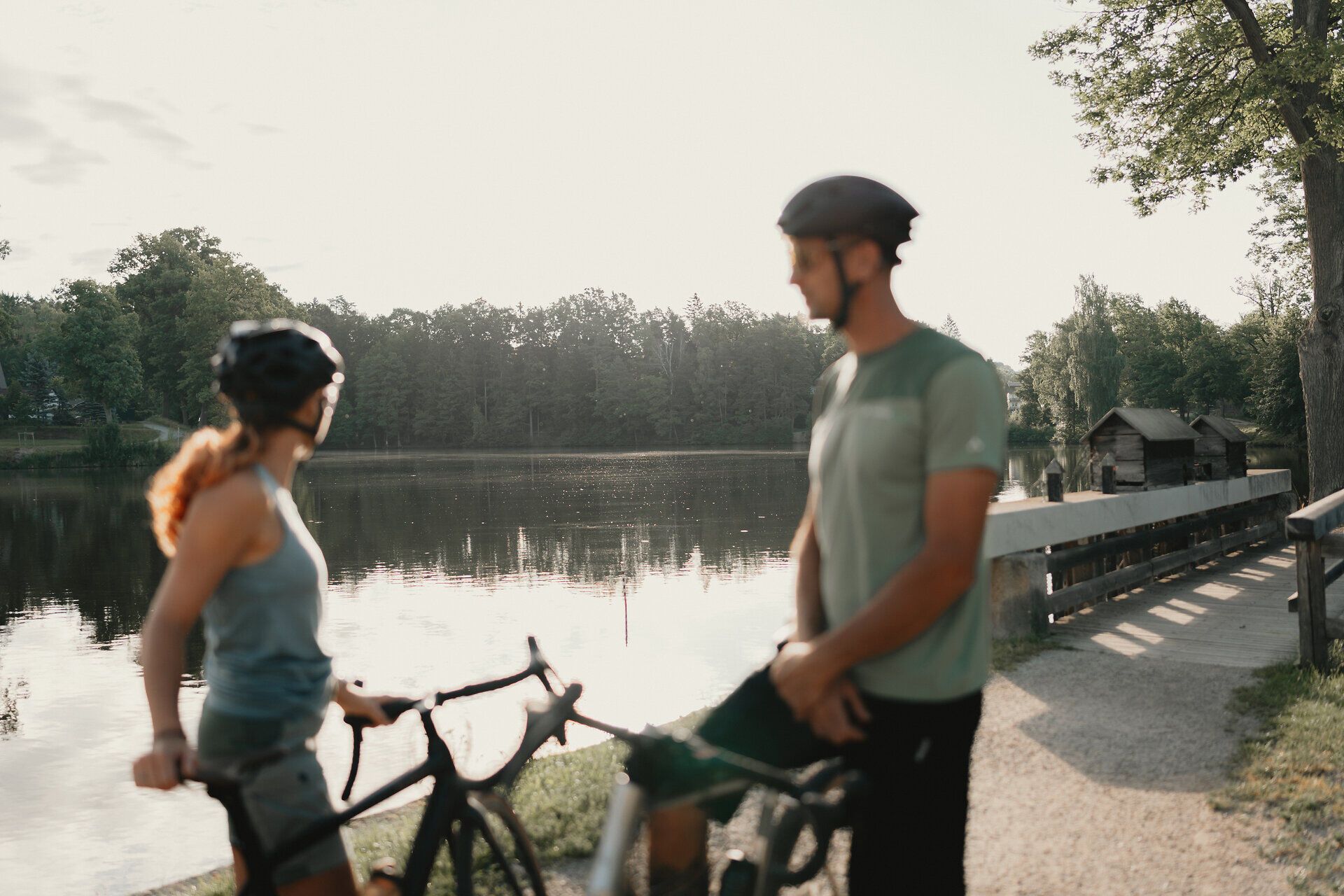 Dva cyklisté v přilbách stojí se svými koly na břehu tichého rybníka obklopeného lesem.