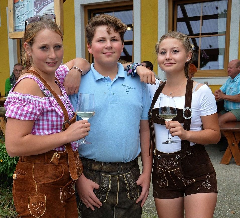 Tři mladí lidé v tradičních bavorských krojích se sklenicemi vína před žlutou budovou.