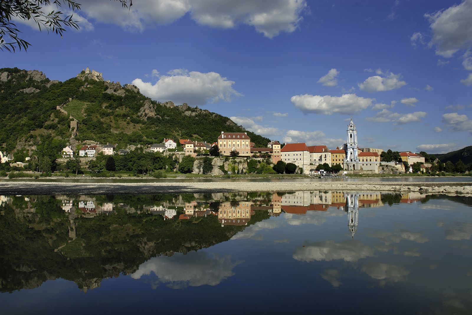Panorama Dürnsteinu s Dunajem v popředí a zříceninou hradu na kopci v pozadí.