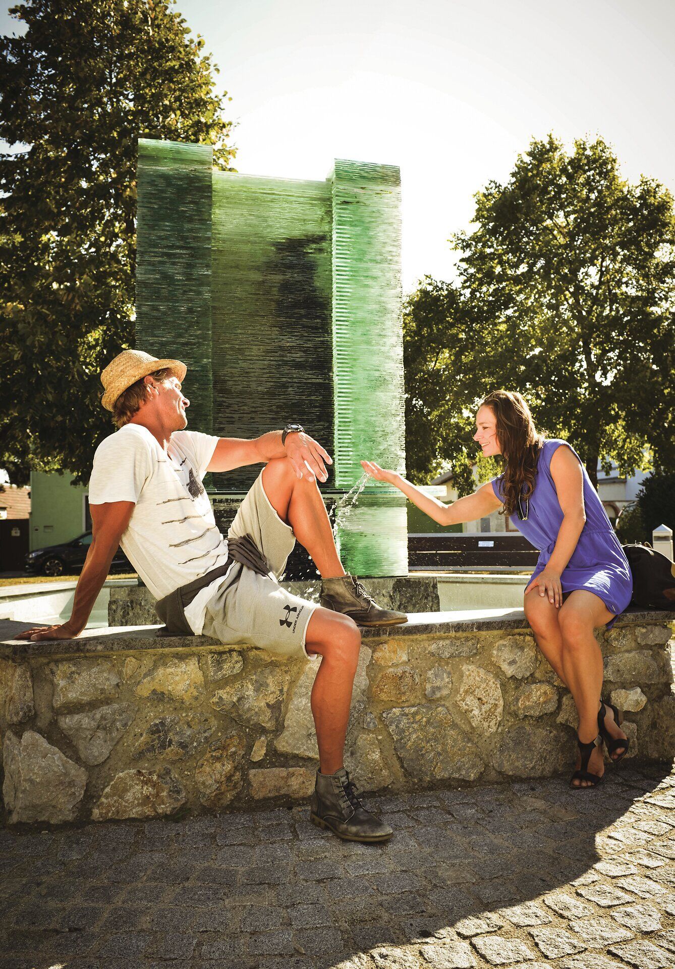 Pár sedí na zdi před fontánou. Žena si hraje s vodou. 