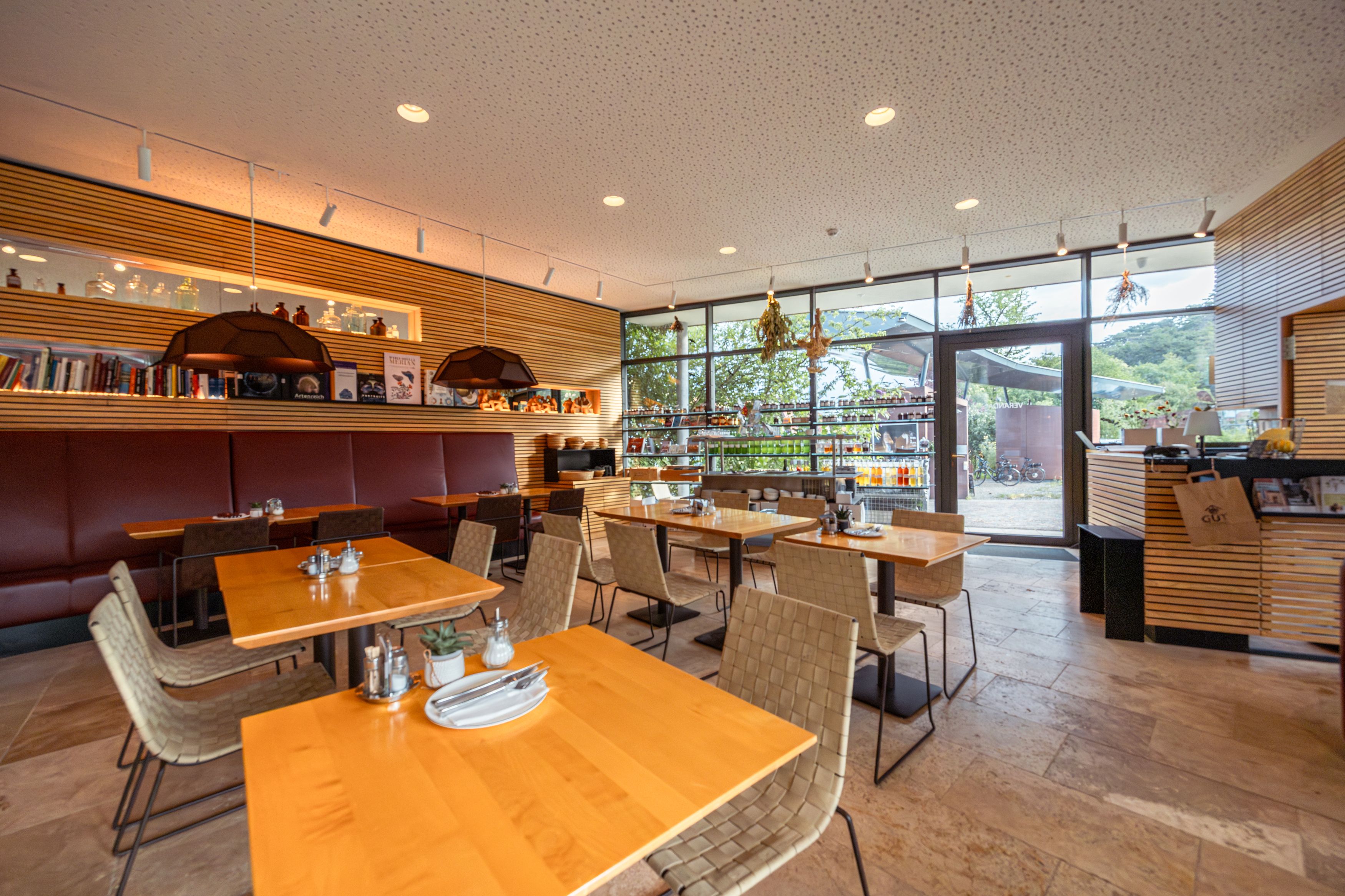 Pohled do interiéru moderní restaurace s dřevěnými stoly a velkými okny.