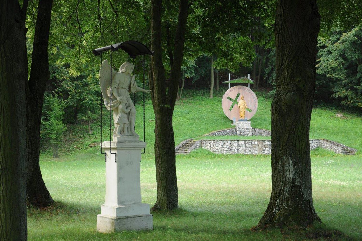 Socha anděla s dítětem v popředí, v pozadí další socha na podstavci v lesoparku.