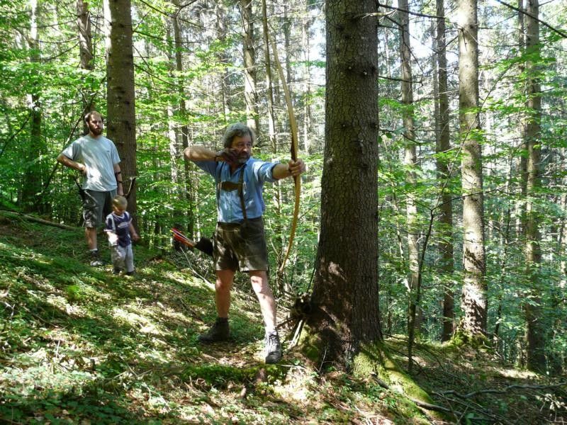 Osoba střílí z luku v lese, v pozadí další dvě osoby.