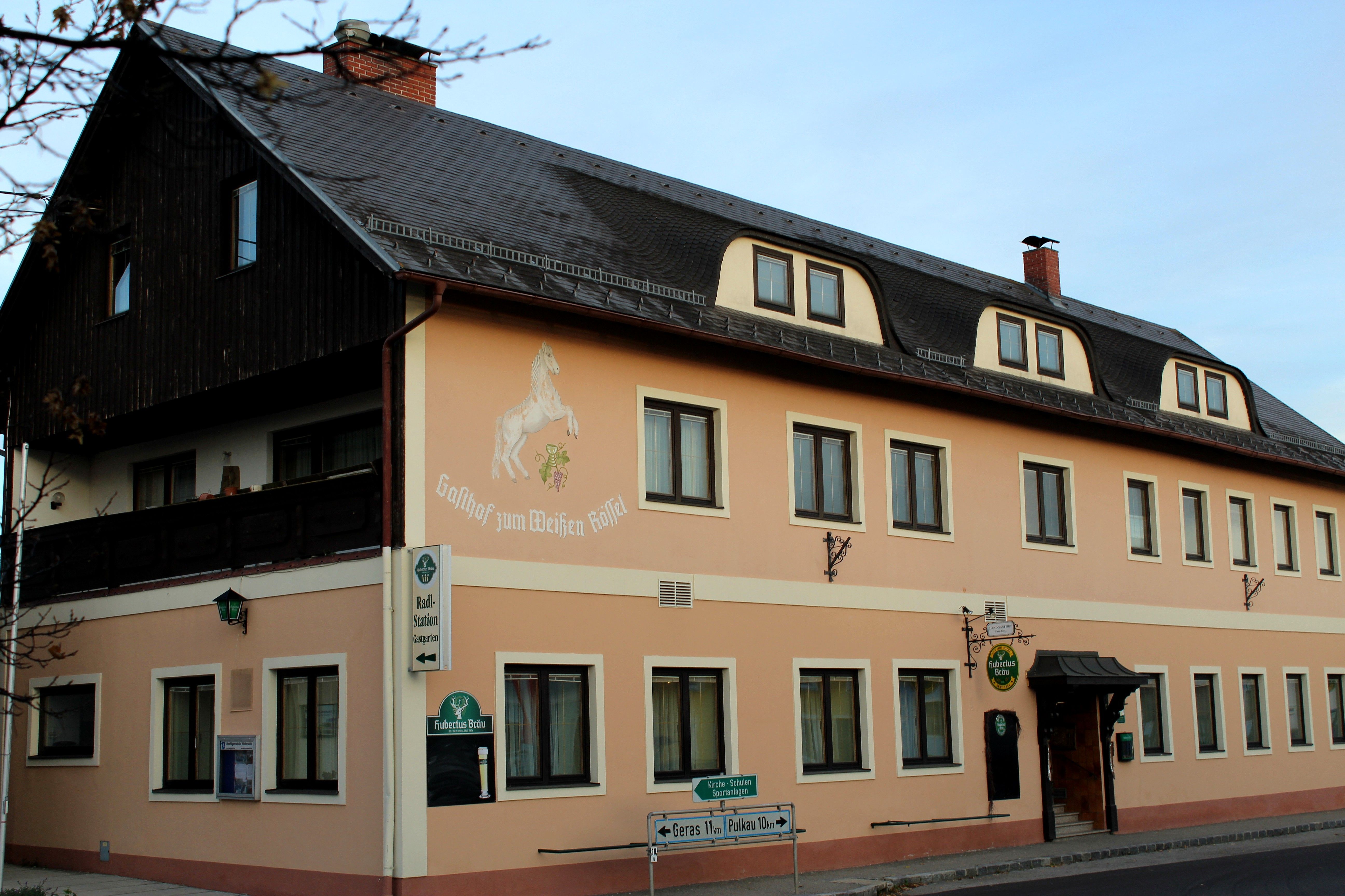 Pohled zvenčí na hostinec "Zum weißen Rössel" s béžovou fasádou a malbou koní.