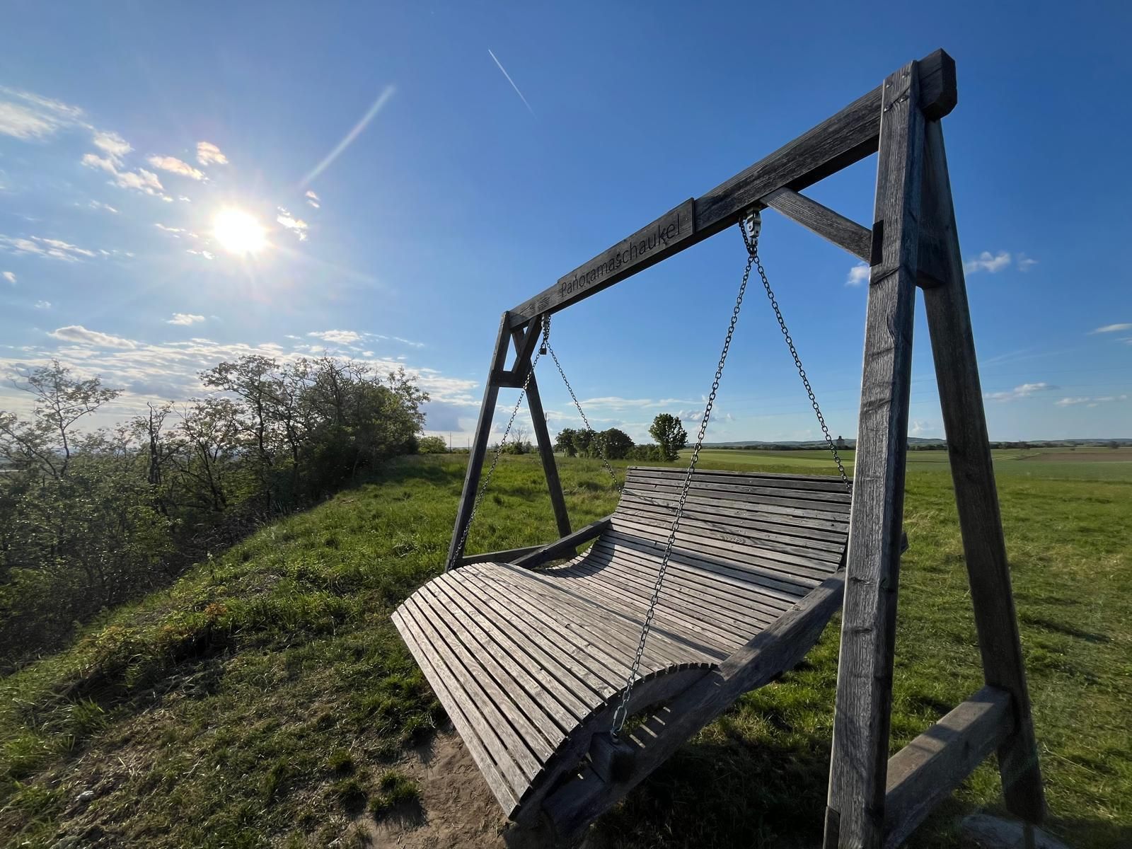 Velká dřevěná panoramatická houpačka na zelené louce pod jasně modrou oblohou. Nízké slunce osvětluje krajinu a vrhá dlouhé stíny, zatímco oblohu křižují jednotlivé mraky a stezka páry. Scéna působí klidně a přívětivě s širokým výhledem na pole a přírodu.