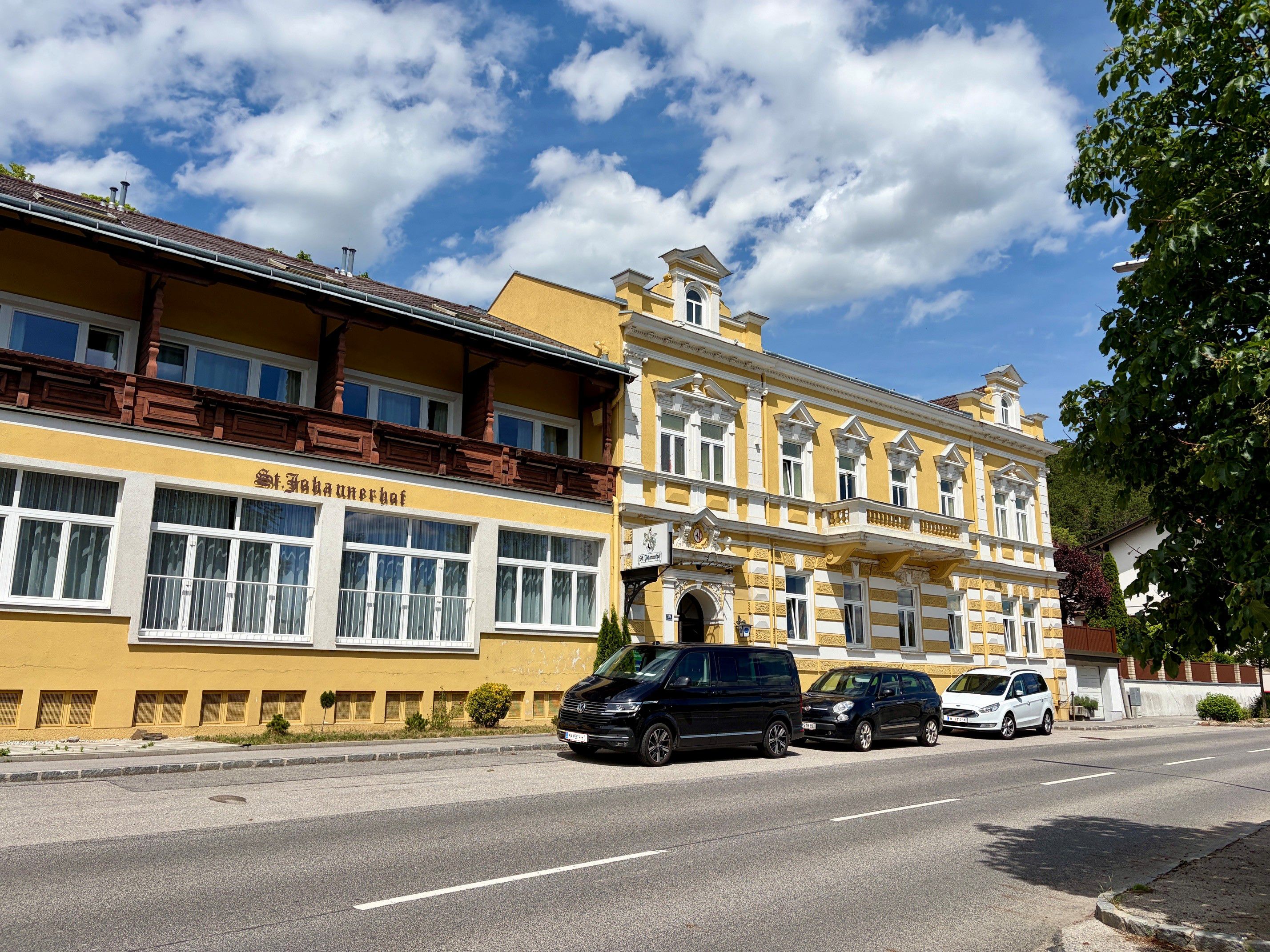 Žlutá budova s nápisem "St. Johannerhof" na silnici, obklopená zaparkovanými auty a stromy.
