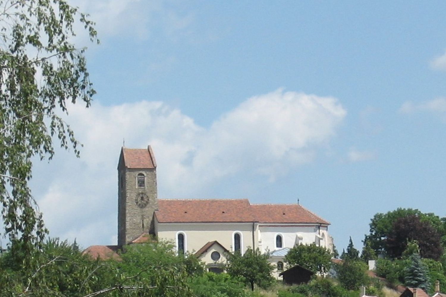 Kostel s věží a červenou střechou v Hohenwarthu, obklopený stromy a modrou oblohou.