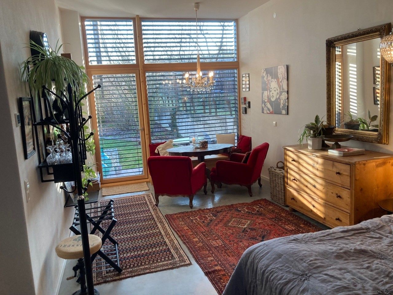 Obývací pokoj s velkými okny, červenými křesly, dřevěným nábytkem a koberci, výhled do zahrady.