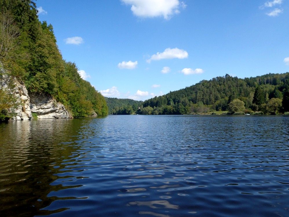 Klidné jezero obklopené zalesněnými kopci pod modrou oblohou s několika mraky.