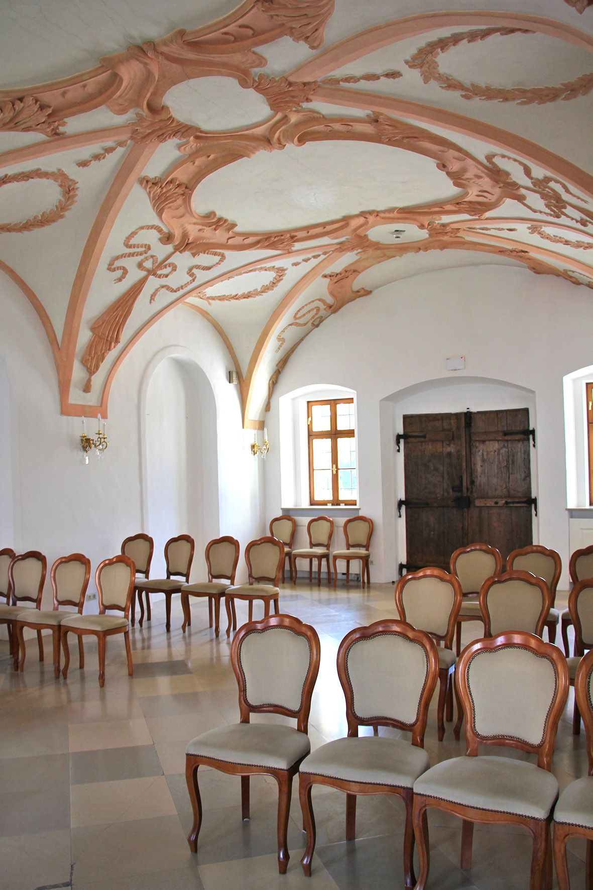 Pohled do interiéru historické místnosti se zdobenými štukovými stropy a elegantními dřevěnými židlemi.