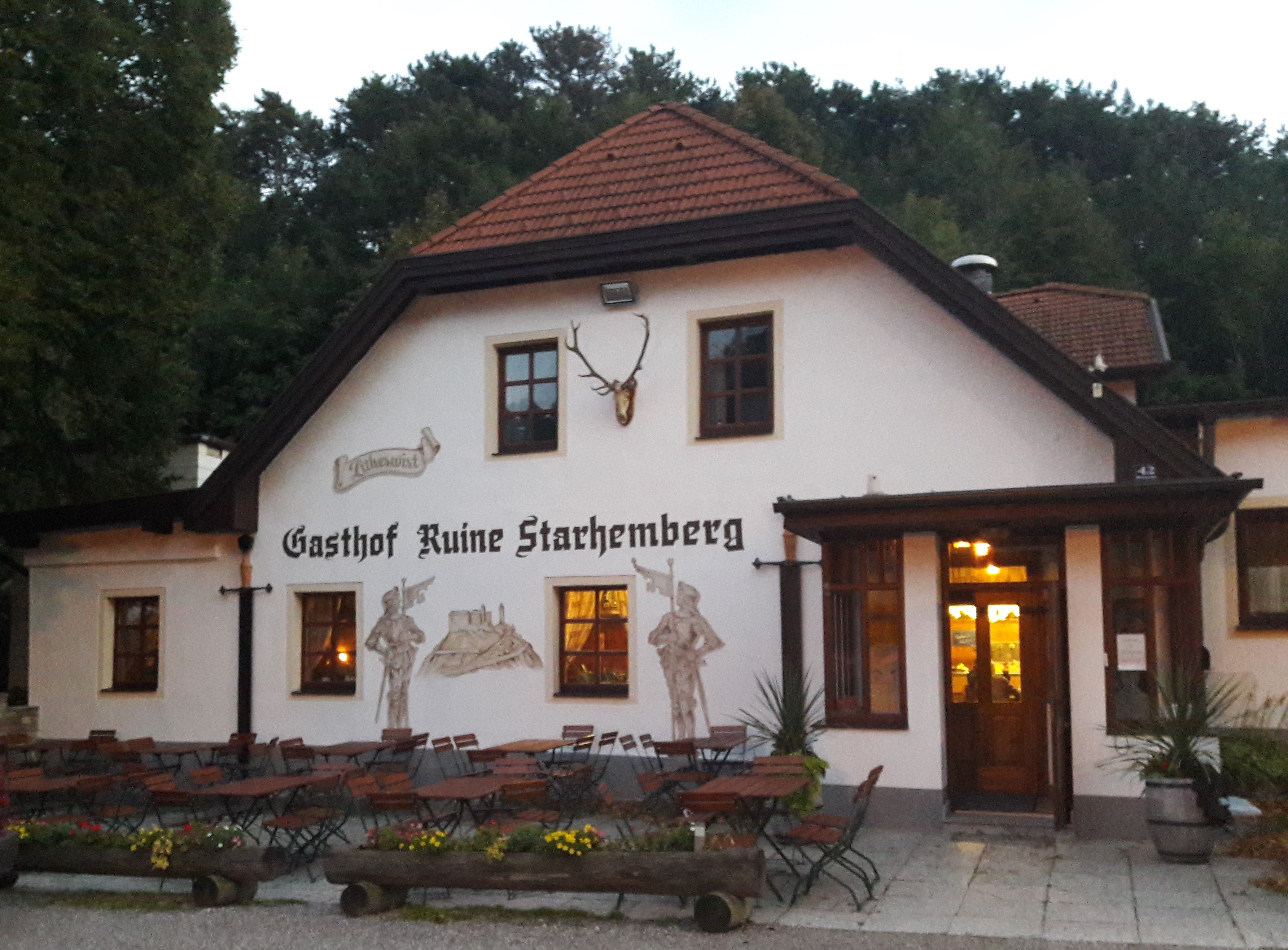 Tradiční hostinec s venkovním prostorem a nástěnnými malbami, obklopený stromy.