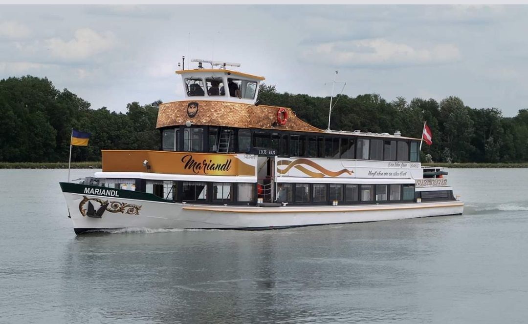Výletní loď MS Mariandl pluje po řece obklopené stromy. Nese vlajky Ukrajiny a Rakouska.