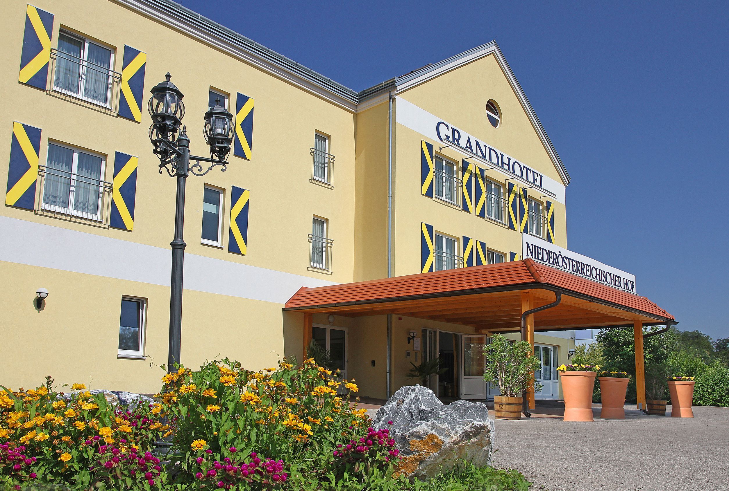 Pohled zvenčí na žlutý hotel s modrými okenicemi a stříškou. Před budovou kvetou žluté a růžové květy.