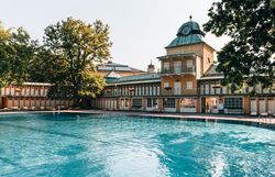 Umgeben von üppigem Grün und majestätischen Bäumen lädt das Thermalbad Vöslau zu erholsamen Stunden ein. Das glitzernde Wasser des Pools reflektiert die warmen Sonnenstrahlen und schafft eine einladende Atmosphäre für Entspannung und Genuss.