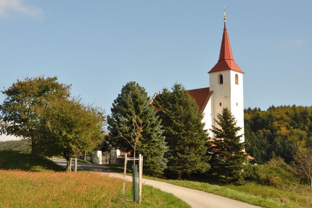 Farní kostel v Ofenbachu s červenou střechou věže, obklopený stromy a loukami.
