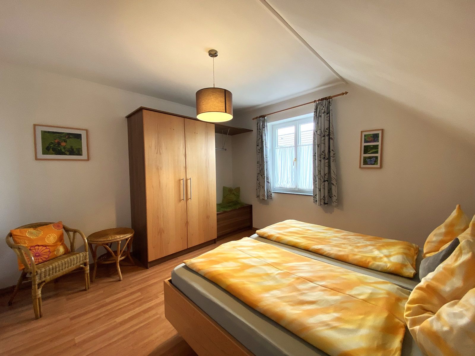 Ložnice s manželskou postelí, dřevěnou skříní, proutěným křeslem a oknem se závěsy.