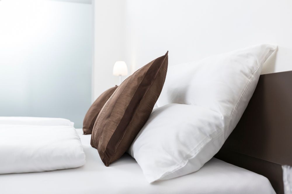 Postel s bílými polštáři a hnědými ozdobnými polštáři, v pozadí lampa.