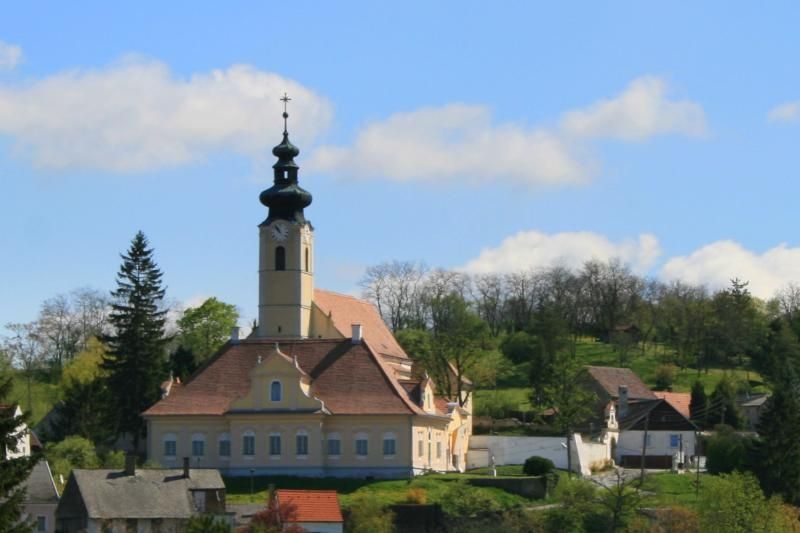 Kostel s věží ve venkovském prostředí, obklopený stromy a domy.
