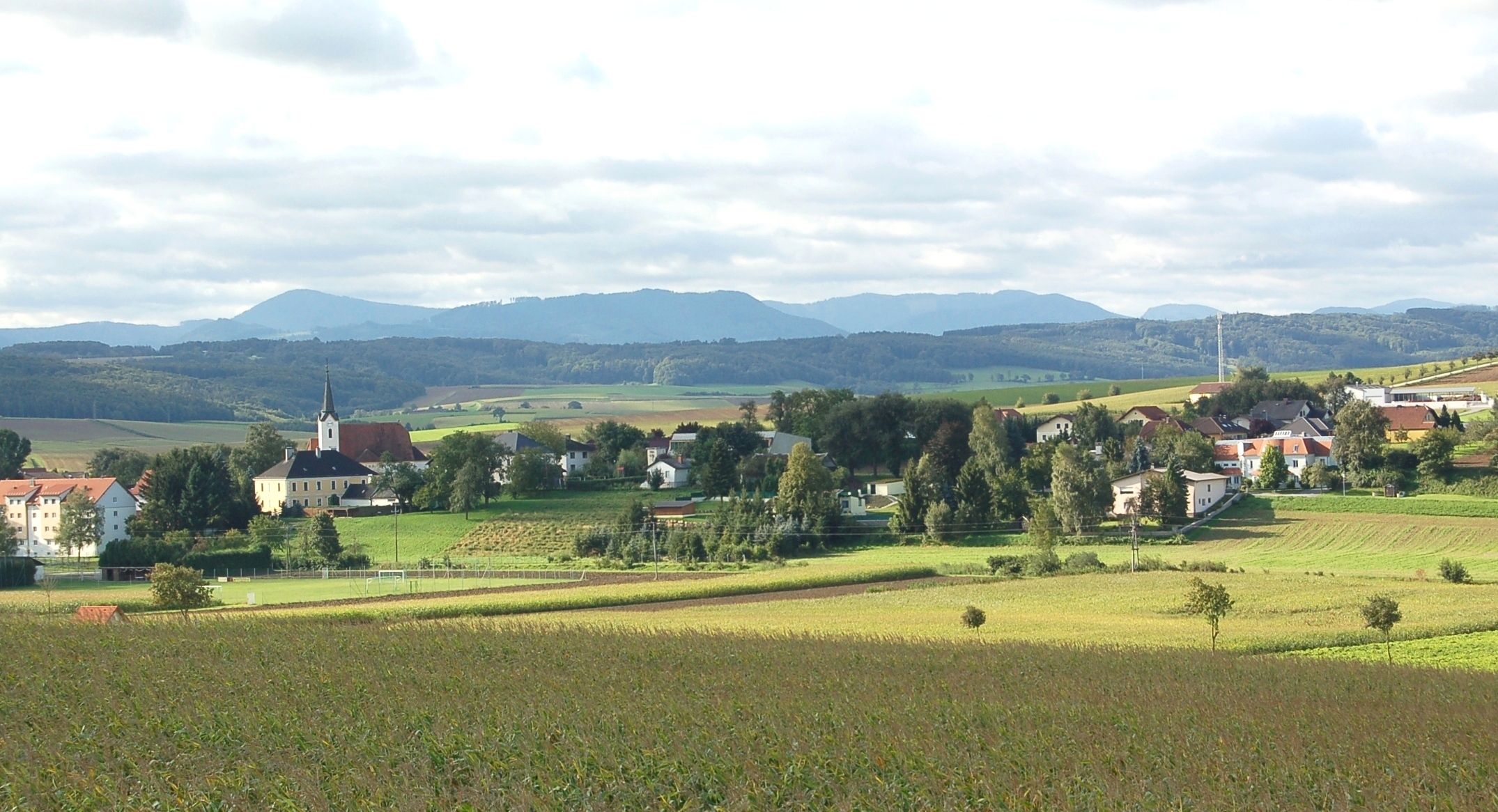 Krajina s vesnicí a kostelem v St. Margarethen an der Sierning, obklopená poli a kopci.