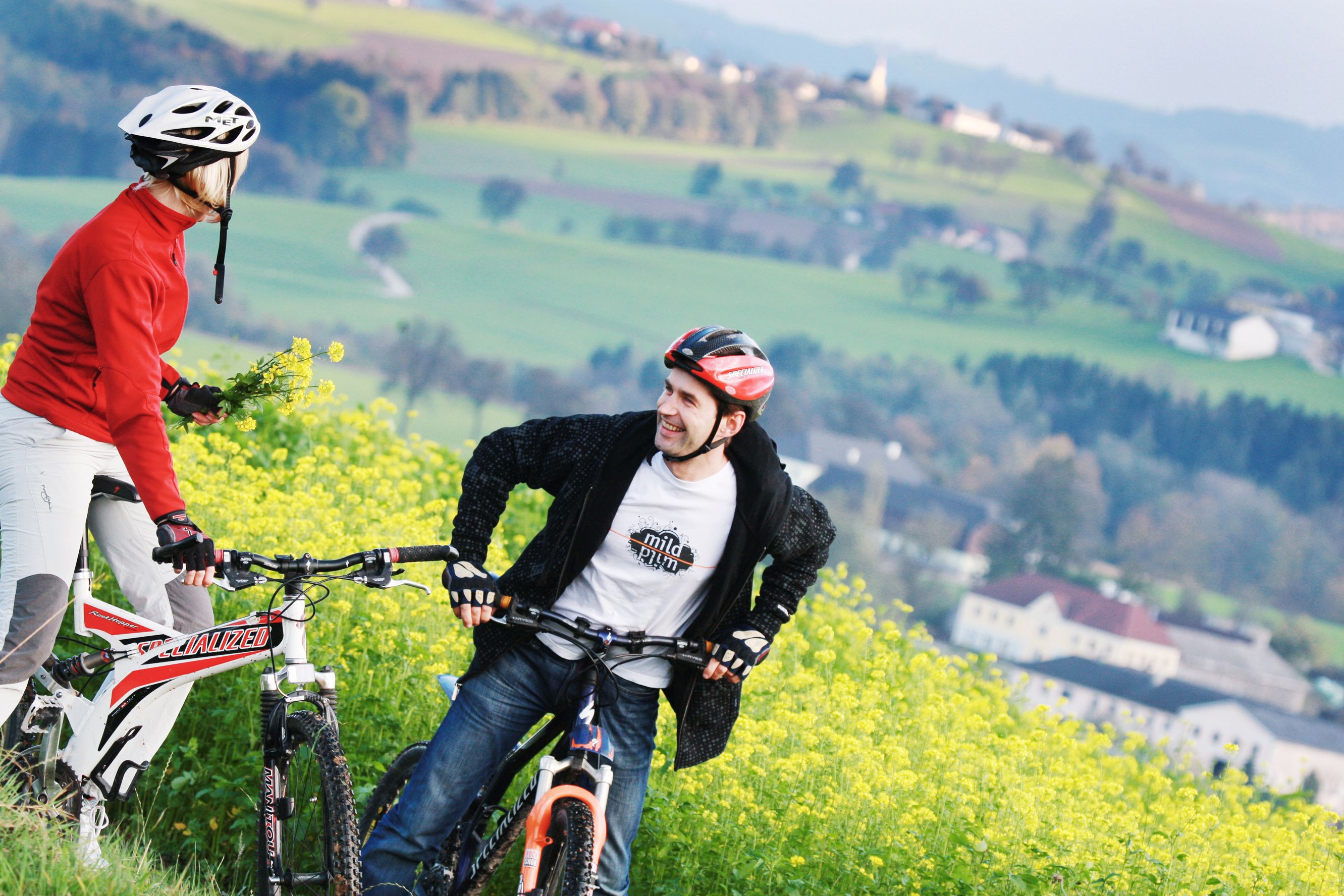 Dva cyklisté s přilbami stojí na rozkvetlém poli se zelenou krajinou v pozadí.