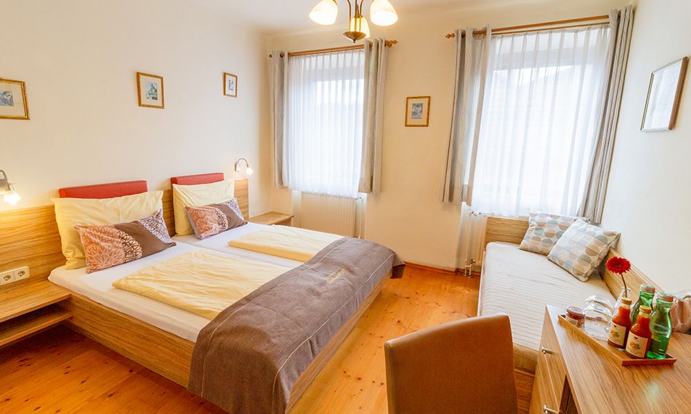Útulný pokoj pro hosty s manželskou postelí, denním lůžkem, dřevěným nábytkem a dekorativními polštáři. Světlé závěsy a obrázky na stěnách.