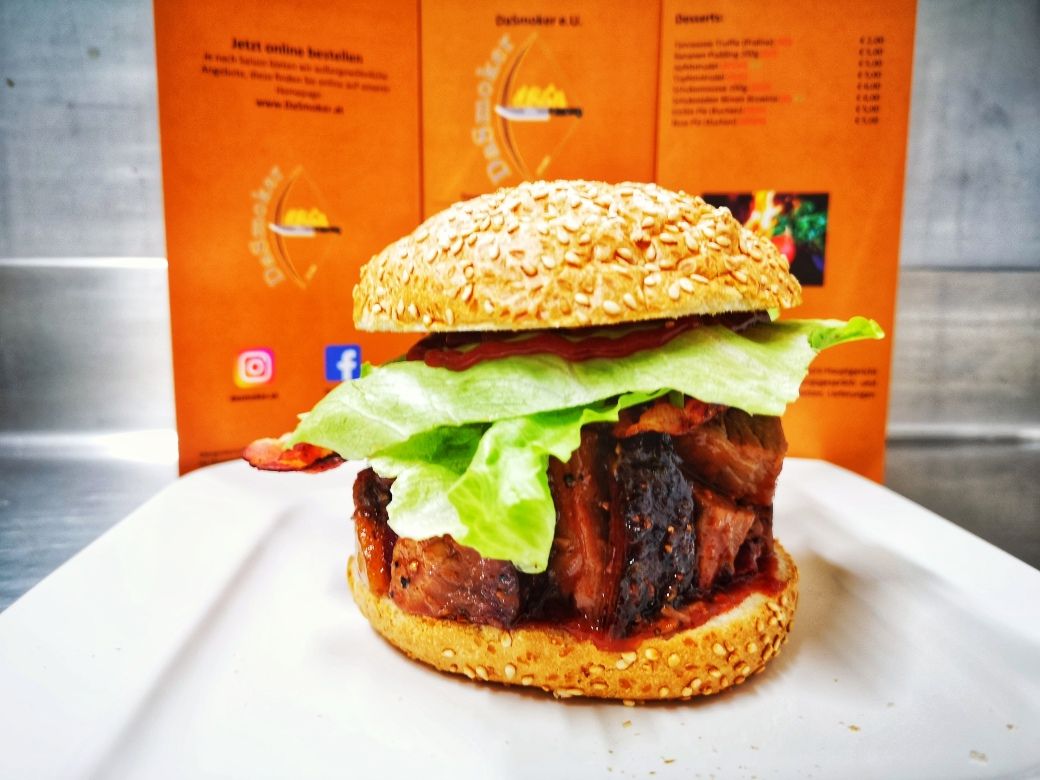 Burger se salátem a masem na bílém talíři před oranžově zbarveným jídelním lístkem.