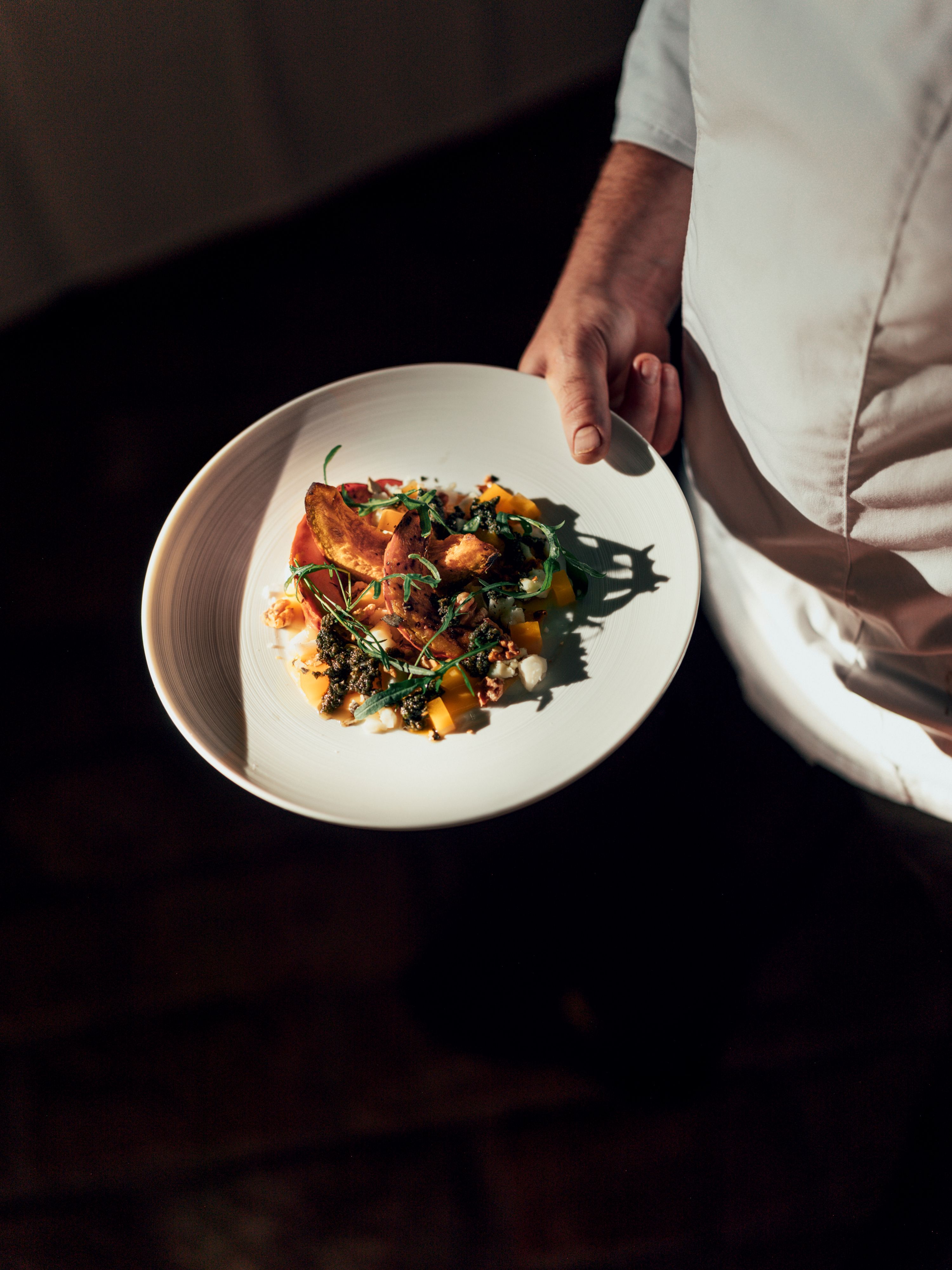 Talíř s pečlivě naaranžovaným pokrmem, který drží osoba v bílém oděvu.