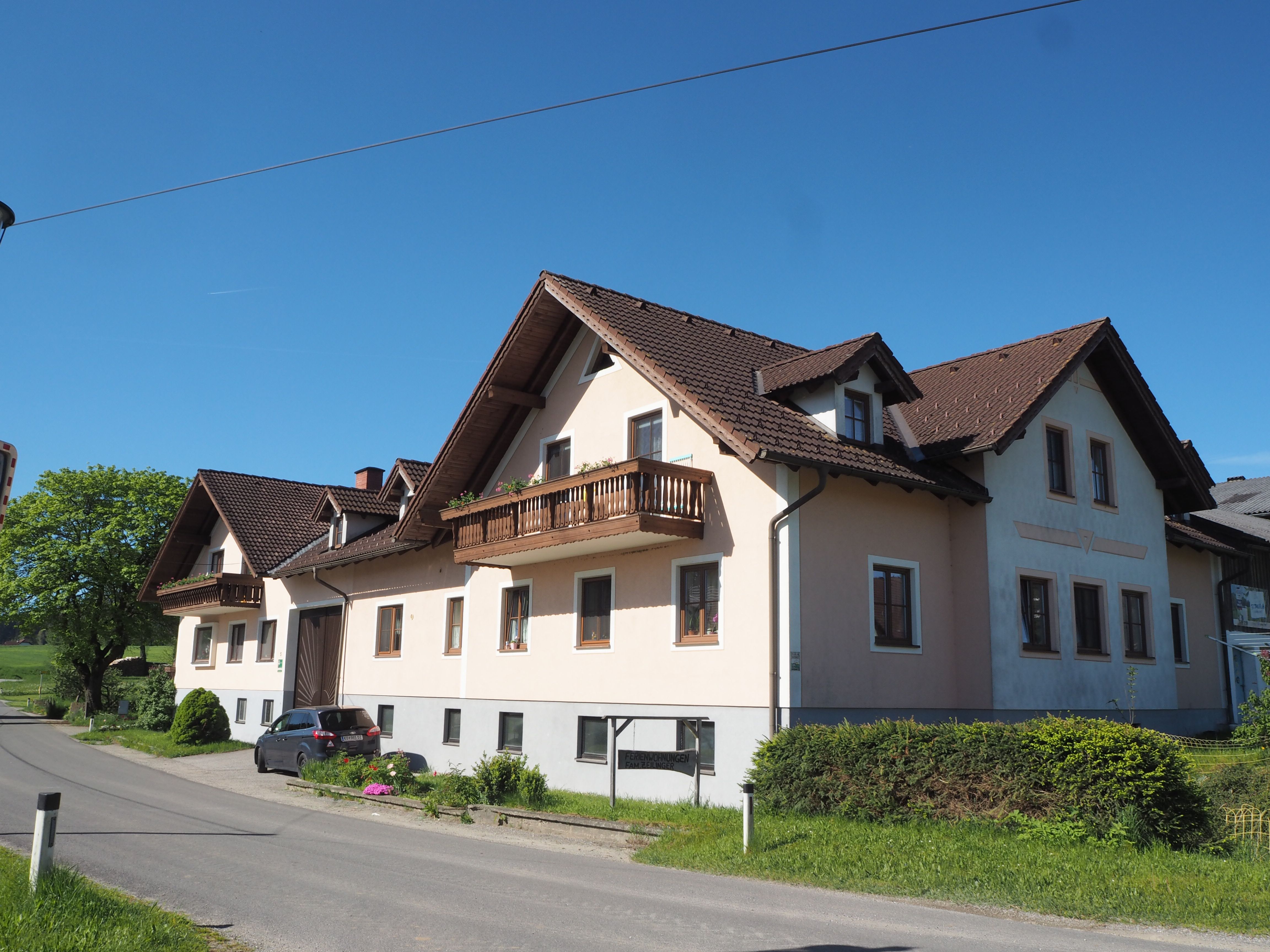 Velký statek s hnědou střechou a balkony na venkovské cestě pod jasnou oblohou.