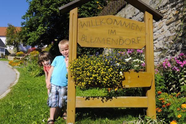 Dvě děti stojí vedle dřevěné cedule s nápisem "Vítejte v květinové vesnici", obklopené barevnými květinami.