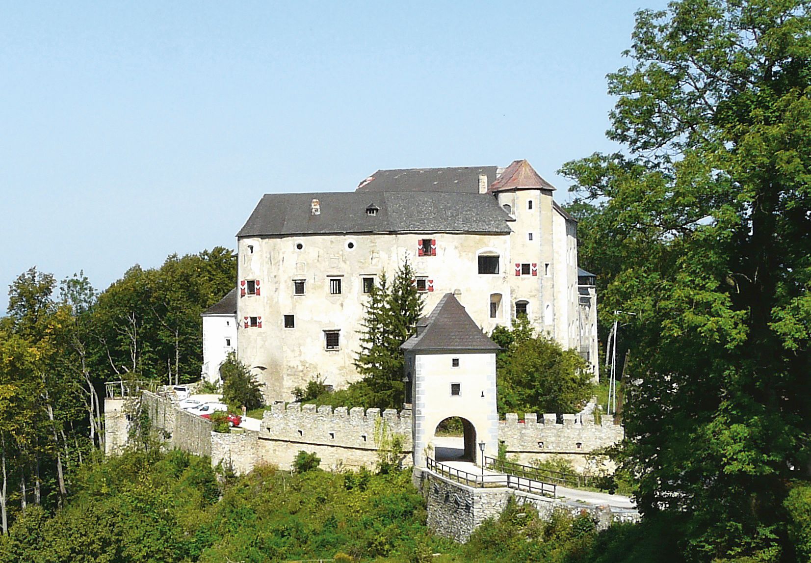 Historický hrad s bílou fasádou a cimbuřím, obklopený stromy.
