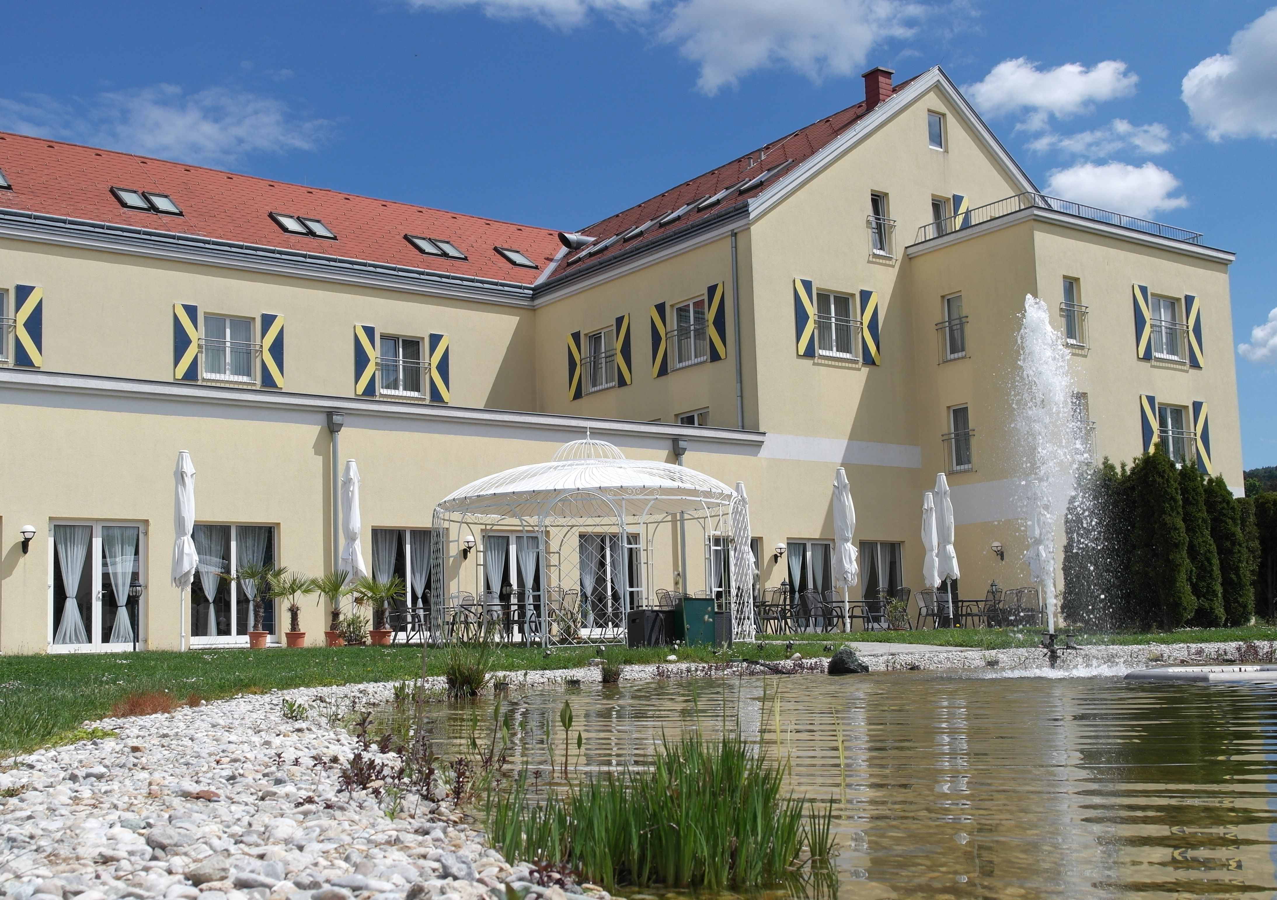 Velká žlutá budova s červenými střešními taškami, fontánou a pavilonem v popředí.