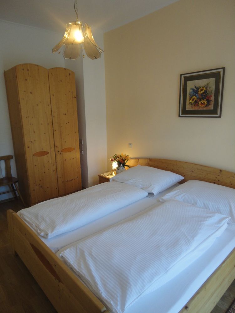 Útulná ložnice s manželskou postelí, dřevěnou skříní, nočním stolkem s květinami a obrazem na stěně.