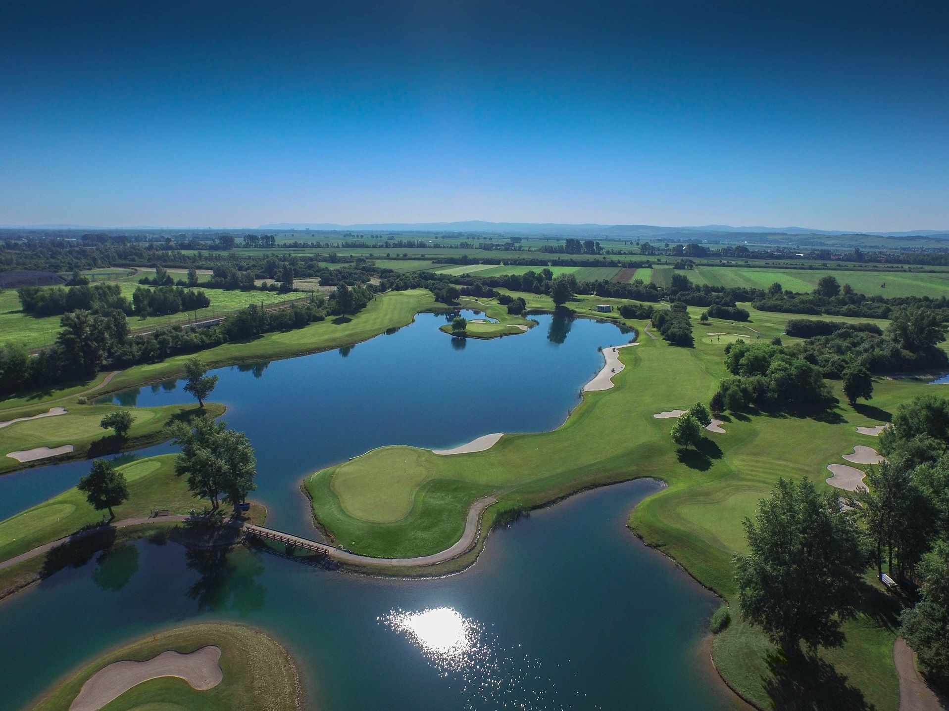 Letecký pohled na golfové hřiště s vodními překážkami a zelenými fairwayemi.