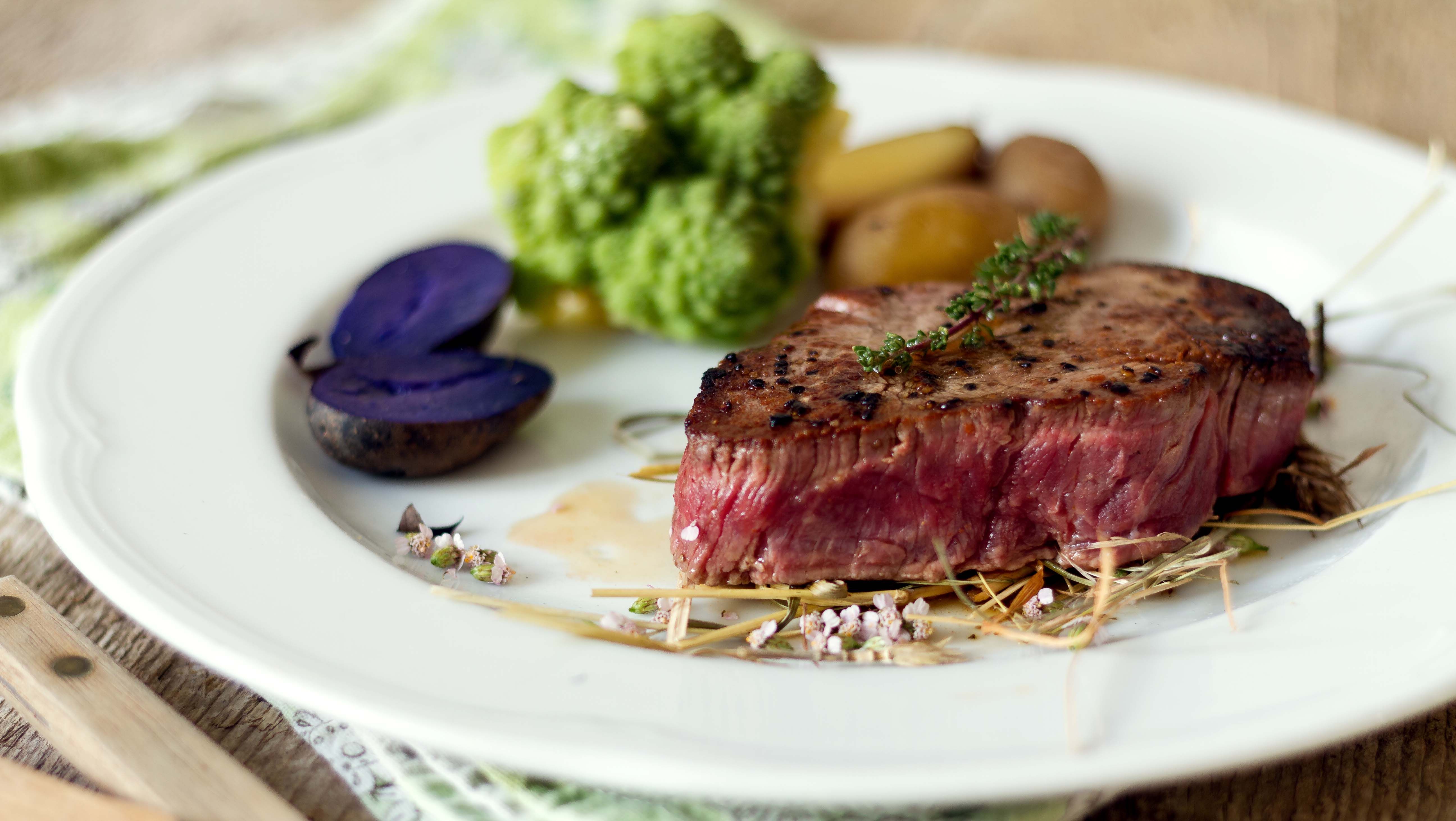 Kousek steaku na bílém kulatém talíři se zeleninou, bramborami a bylinkami.
