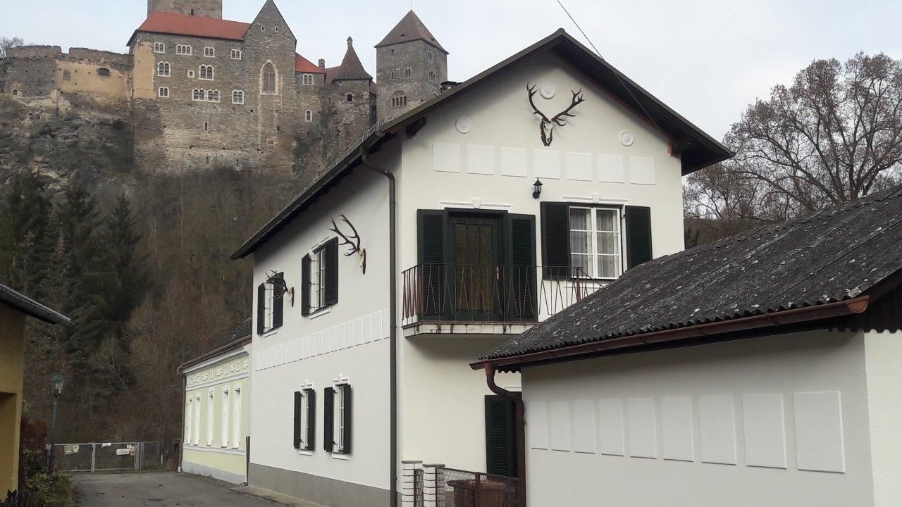 Bílý dům se zelenými okenicemi a jeleními parohy na zdi, v pozadí hrad na kopci.