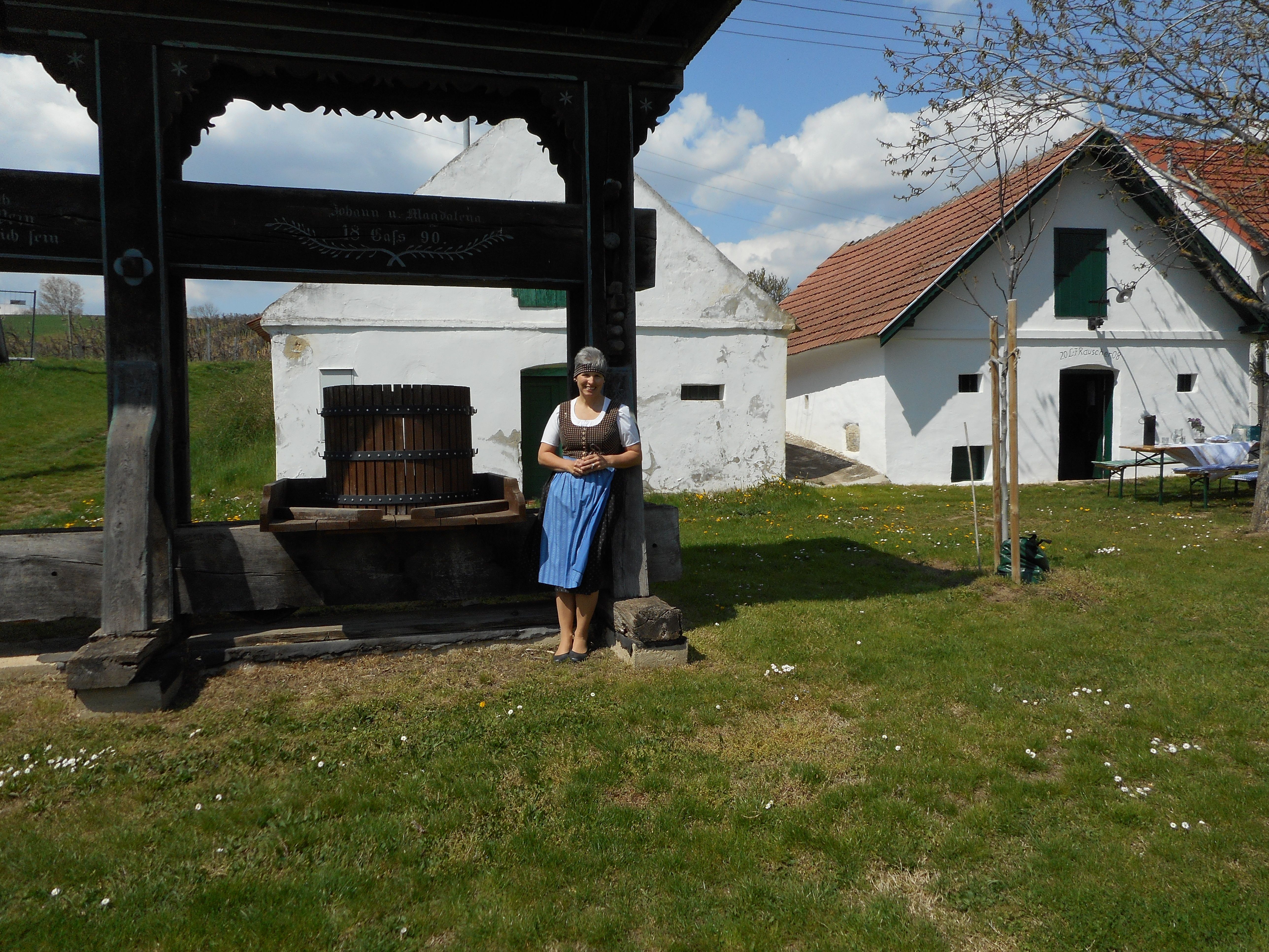 Žena v tradičním oděvu stojí vedle starého lisu na víno před historickou budovou s bílými zdmi a červenými střechami.