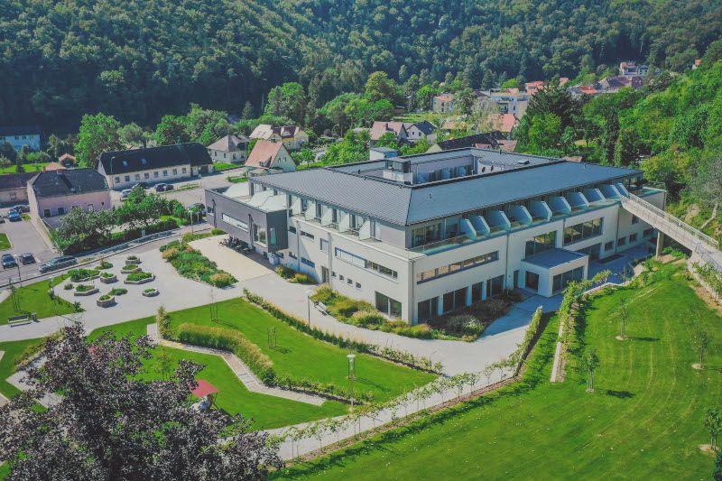 Letecký pohled na moderní budovu se zahradou a okolní krajinou.